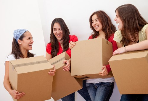 Four young women with light skin and long hair are indoors, possibly in a home or apartment, engaged in a moving or packing activity. They are smiling and appear to be in good spirits. Two of the women, on the far left and second from the right, are holding medium-sized cardboard boxes, with one woman wearing a light blue headscarf and a white top, and the other wearing a red top and blue jeans. The woman on the far right, dressed in a yellow top, is leaning on a large cardboard box, partially obscured. In the background, the wall is plain white with no visible furniture or decorations, indicating a clean, uncluttered interior space. The scene captures a moment of teamwork associated with house removals or relocation services, relevant to companies operating in Richmond upon Thames. The natural indoor lighting highlights the cardboard boxes’ brown color and the different textures of the clothing, emphasizing a friendly and professional atmosphere suitable for a house moving service web page.
