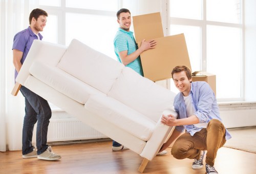 Three male movers are inside a well-lit room with large windows, engaged in carrying and positioning a light beige fabric-covered sofa with wooden legs. The mover on the left, dressed in a purple T-shirt, is holding the left side of the sofa, while the mover on the right, wearing a light blue button-up shirt and brown trousers, is crouching down with his hands on the sofa's bottom edge, stabilising it. The third mover, standing behind and slightly to the right,, wearing a turquoise T-shirt, is holding a medium-sized, plain cardboard box, preparing to pass or move it. The room has a wooden floor, a white radiator beneath the windows, and no furniture or other objects visible, providing a clean, minimal background. The scene depicts the process of a residential move, typical of house removals, within the context of a property in Richmond upon Thames, with natural light illuminating the space, and the focus on the handling of furniture and packing items as part of the relocation task carried out by [COMPANY_NAME].
