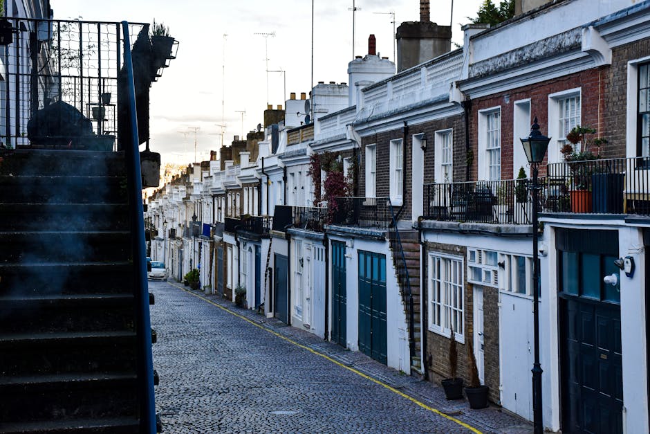 A narrow residential street with a cobblestone surface in Richmond, showing a row of terraced houses with brick and white-painted facades, some featuring small front gardens with potted plants and dec