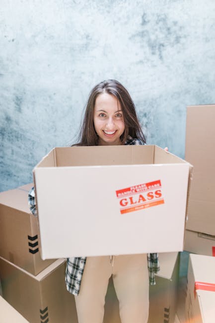 A woman with long brown hair and a smile is holding an empty cardboard box labeled 'FRAGILE GLASS' inside a room with a textured light blue wall. She is standing near other packed boxes, some with bla