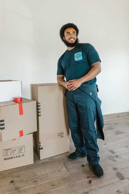 A woman with long brown hair and a smile is holding an empty cardboard box labeled 'FRAGILE GLASS' inside a room with a textured light blue wall. She is standing near other packed boxes, some with bla